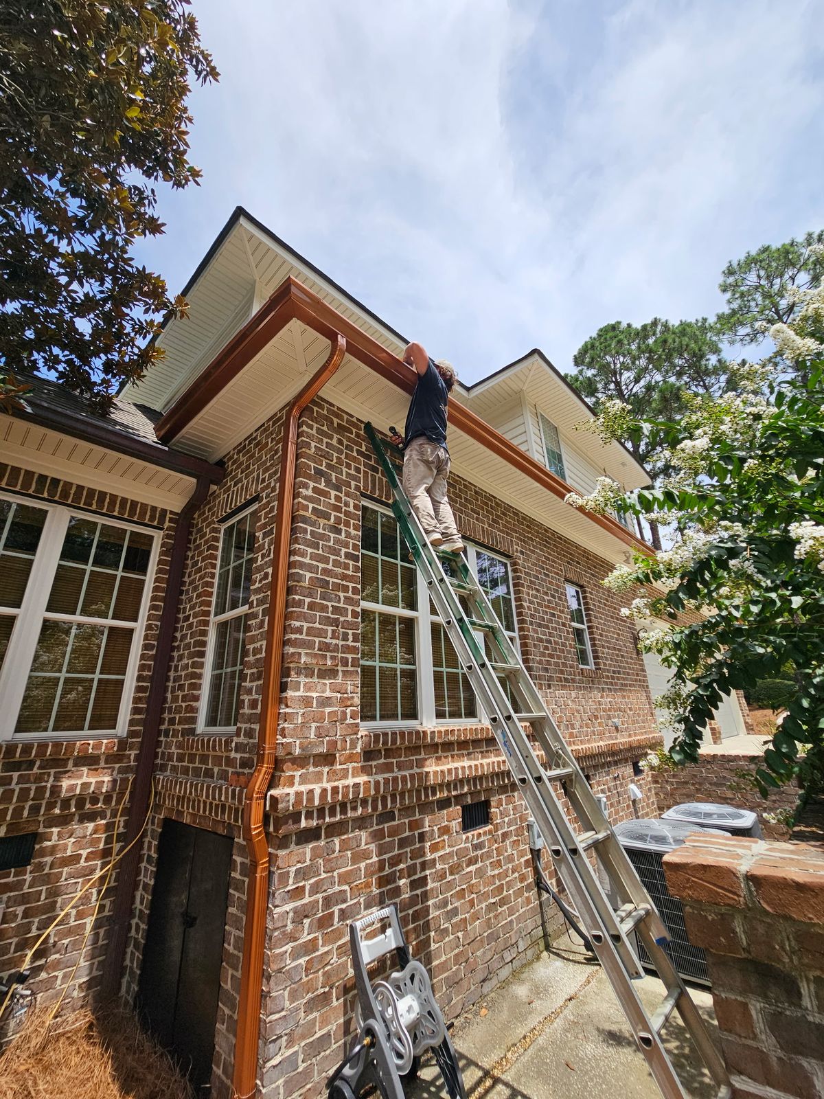 Installer on a ladder fitting copper-colored gutters and a downspout on a two-story brick house