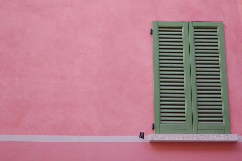Pair of green louvered exterior shutters on a window in a pink stucco wall